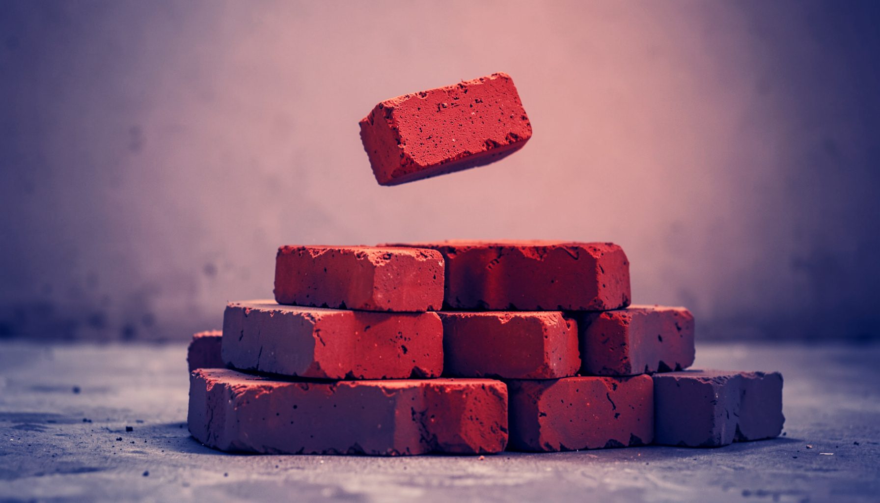 Duotone photograph of stacked red bricks on a concrete floor in soft navy and coral tones, a single brick mid-air above